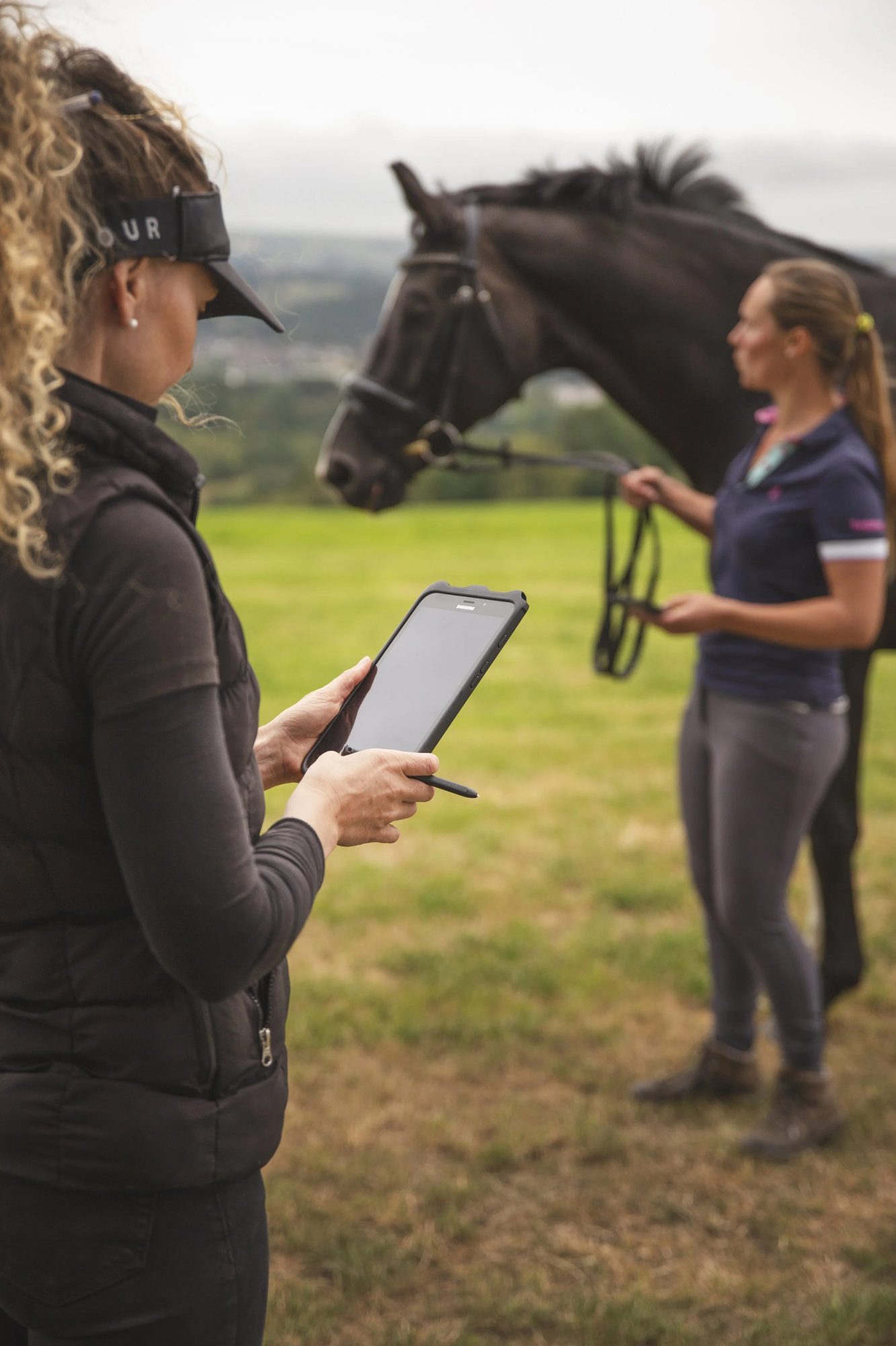 An equine therapist in a field using the Equigate app on a tablet while a client holds her horse